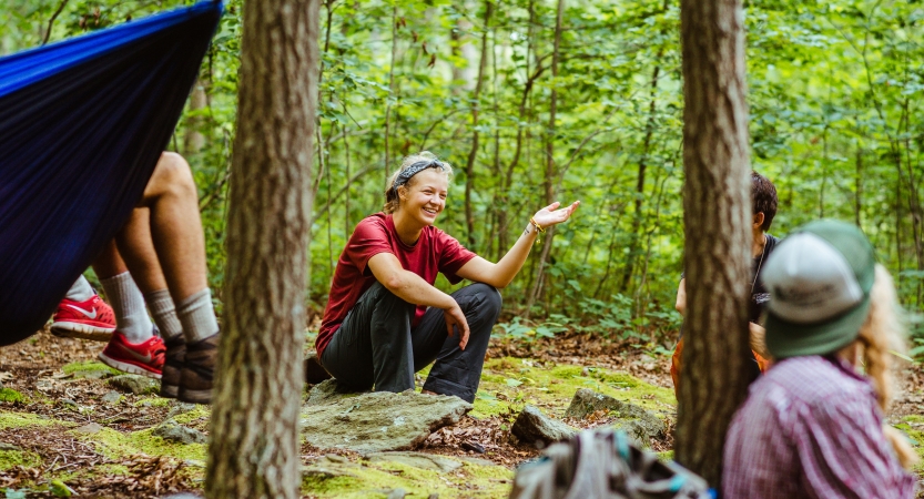 A person smiles while sitting on the ground in a wooded area. You can see the legs of two people resting in a hammock and others on the ground, too. 